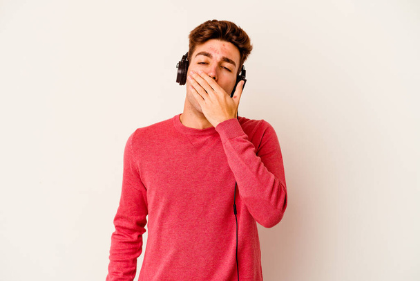 Молодий кавказький чоловік слухає музику ізольовано на білому тлі позіхання, показуючи втомлений жест, що покриває рот рукою
. - Фото, зображення