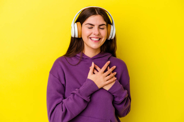 Young caucasian woman listening to music with headphones isolated on pink background laughing keeping hands on heart, concept of happiness. - Valokuva, kuva