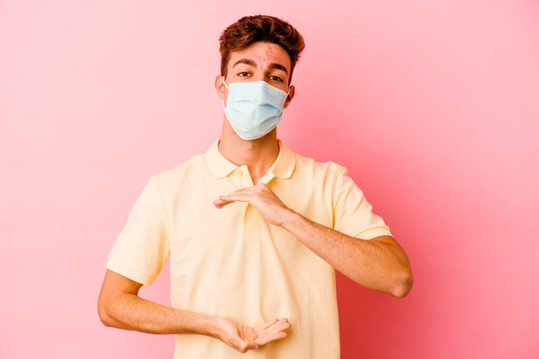 Joven hombre caucásico usando una protección para coronavirus aislado sobre fondo rosa sosteniendo algo con ambas manos, presentación del producto. - Foto, Imagen