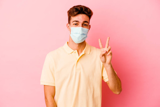 Jeune homme caucasien portant une protection contre le coronavirus isolé sur fond rose montrant signe de victoire et souriant largement. - Photo, image