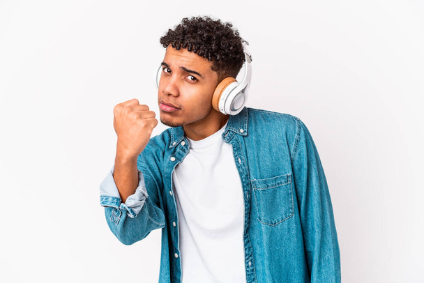 Joven afroamericano rizado hombre aislado escuchando música con auriculares mostrando puño a cámara, expresión facial agresiva. - Foto, Imagen