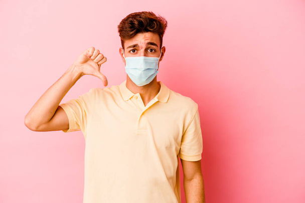 Hombre caucásico joven que lleva una protección para el coronavirus aislado sobre fondo rosa que muestra un gesto de aversión, pulgares hacia abajo. Concepto de desacuerdo. - Foto, Imagen