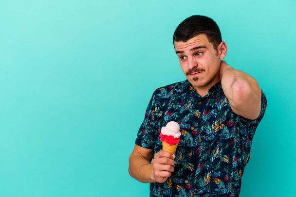 頭の後ろに触れる青の背景に隔離されたアイスクリームを食べている若い白人男性、思考と選択をする. - 写真・画像