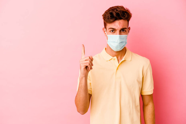 Jeune homme caucasien portant une protection contre le coronavirus isolé sur fond rose montrant numéro un avec doigt. - Photo, image