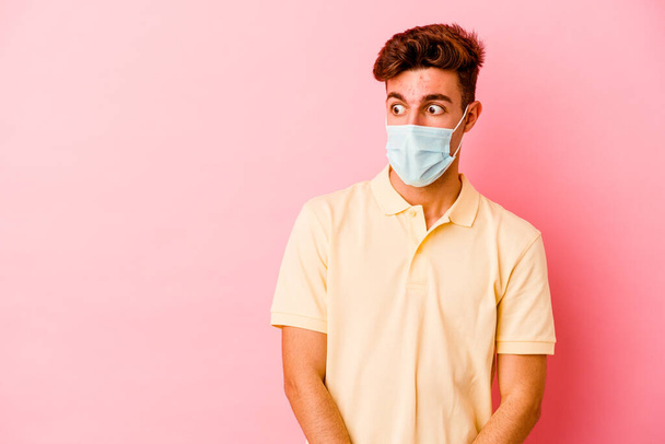 Young caucasian man wearing a protection for coronavirus isolated on pink background being shocked because of something she has seen. - Photo, Image