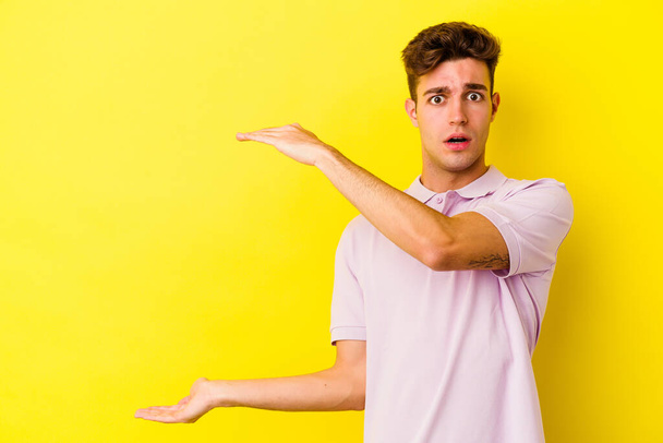 Young caucasian man isolated on yellow background shocked and amazed holding a copy space between hands. - Photo, Image