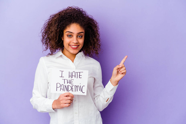Молодая афроамериканка держит я ненавижу пандемический плакат изолированный на фиолетовом фоне улыбаясь и указывая в сторону, показывая что-то в пустом пространстве. - Фото, изображение