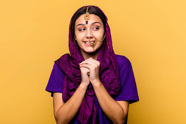 Young Indian woman wearing a traditional sari clothes isolated on yellow background keeps hands under chin, is looking happily aside. - Foto, Imagen