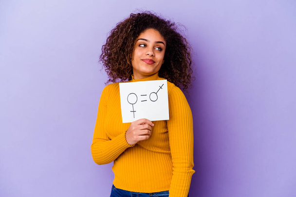 Jonge Afro-Amerikaanse vrouw met een gendergelijkheid bordje geïsoleerd op paarse achtergrond kijkt opzij glimlachend, vrolijk en aangenaam. - Foto, afbeelding