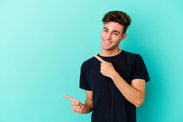 Homem caucasiano jovem isolado em fundo azul apontando com os dedos indicador para um espaço de cópia, expressando emoção e desejo. - Foto, Imagem