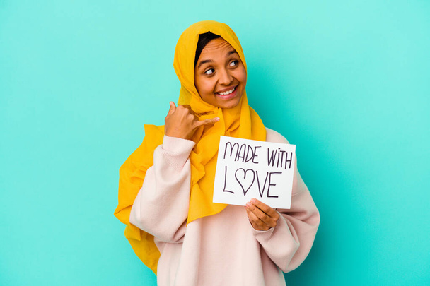 Νεαρή μουσουλμάνα γυναίκα κρατώντας ένα κατασκευασμένο με αγάπη πλακάτ που απομονώνονται σε μπλε φόντο δείχνει μια χειρονομία κινητής τηλεφωνίας με τα δάχτυλα. - Φωτογραφία, εικόνα