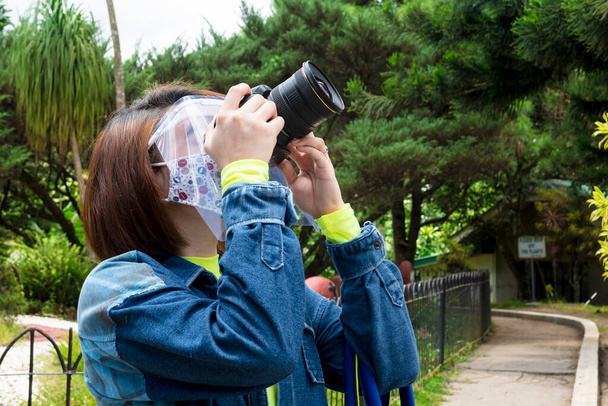 Een Aziatische vrouw met een gezichtsmasker en schild probeert een foto te maken met een SLR-camera. - Foto, afbeelding