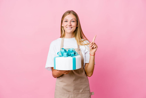 Young russian baker woman holding a delicious cake indicates with both fore fingers up showing a blank space. - Foto, afbeelding