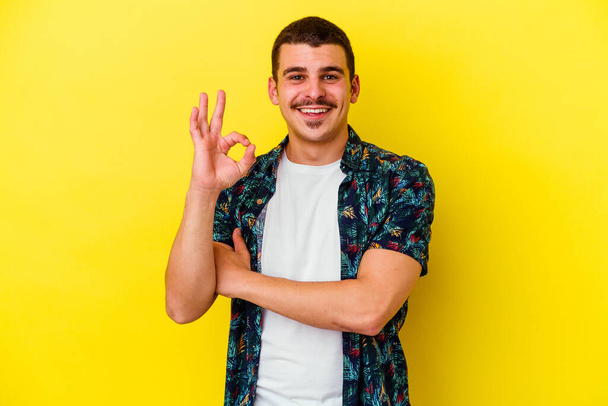 Молодой кавказский холодный человек, изолированный на жёлтом фоне, подмигивает глазом и держит нормальный жест рукой.. - Фото, изображение