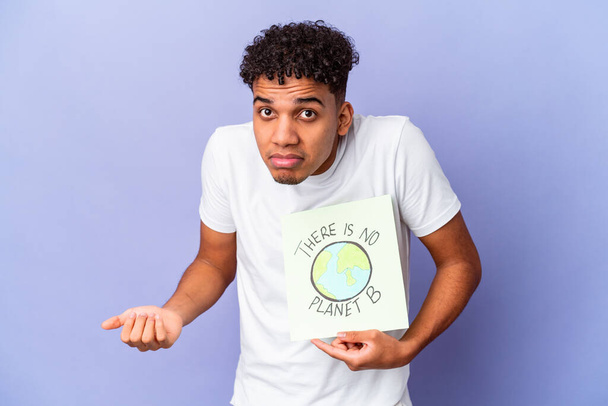 Young african american curly man isolated holding a theres is no planet b poster shrugs shoulders and open eyes confused. - Photo, Image
