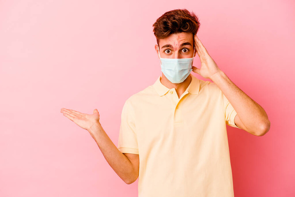 Junger kaukasischer Mann mit einem Schutz gegen Coronavirus isoliert auf rosa Hintergrund hält Kopierraum auf einer Handfläche, die Hand über die Wange. Erstaunt und erfreut. - Foto, Bild