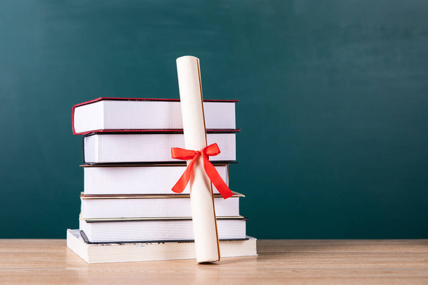 Kırmızı kurdeleli bir diploma ve yeşil karatahta arkaplan üzerine bir yığın kitap. - Fotoğraf, Görsel