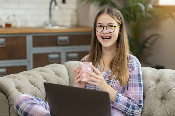 Happy teenager with a smile in pajamas and a laptop holds a cup of tea and looks directly into the camera while sitting on the couch at home - Φωτογραφία, εικόνα
