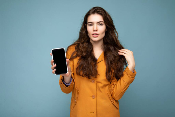sexy piękny młody brunetka kobieta noszenie pomarańczowy kurtka odizolowany nad niebieski tło trzyma w dłoni i pokazując telefon komórkowy z pusty wyświetlacz dla makieta patrząc w aparat - Zdjęcie, obraz