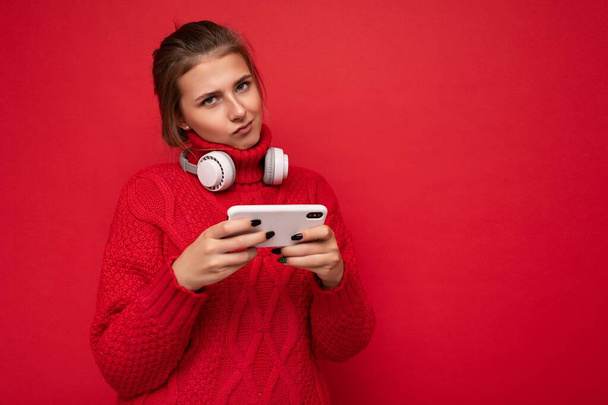 Kırmızı arka plan duvarından izole edilmiş kırmızı süveter giyen çekici genç esmer kadın internette cep telefonu kullanıyor ve bluetooth kulaklık takıyor. - Fotoğraf, Görsel