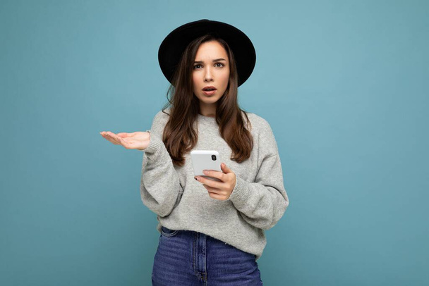 Красива молода дівчина питає незадоволену брюнетку в чорному капелюсі і сірий светр тримає смартфон, дивлячись на камеру ізольовано на фоні
 - Фото, зображення