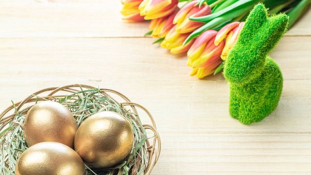Πασχαλινά αυγά χρυσά. Χρυσό χρώμα αυγό σε καλάθι με ανοιξιάτικες τουλίπες, λευκά φτερά σε ξύλινο φόντο σε Happy Easter διακόσμηση. Σχεδιασμός επετηρίδων συγχαρητηρίων - Φωτογραφία, εικόνα