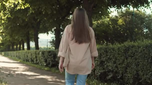 Παρακολούθηση πίσω όψη του ευτυχισμένη νεαρή Καυκάσια γυναίκα περπάτημα άλμα στο ηλιόλουστο καλοκαιρινό πάρκο, στη συνέχεια, γυρίζοντας γύρω, κουνώντας το χέρι, χαμογελώντας και κοιτάζοντας την κάμερα - Πλάνα, βίντεο