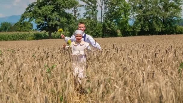 Dvě děti skákají přes pšeničné pole a drží slunečnice. Pomalý pohyb - Záběry, video