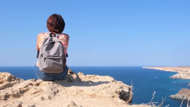 Jonge gelukkige vrouw met een rugzak zit op een rots en kijkt naar de vallei beneden. Toeristen aan de rotskust van de Zwarte Zee. Het concept van vrijheid en gemak - Video