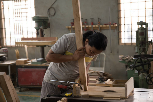 Ένας νεαρός άνδρας ξυλουργός οικοδόμος σε ένα γκρι T-shirt και εργασίας συνολικά ισούται με ένα ξύλινο μπαρ με μια μηχανή άλεσης στο εργαστήριο, στο φόντο ξύλινες σανίδες, στο εργαστήριο - Φωτογραφία, εικόνα