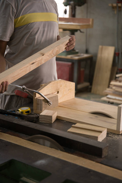 Un joven carpintero constructor masculino con una camiseta gris y trabajando en general equivale a una barra de madera con una fresadora en el taller, en las tablas de madera de fondo, en el taller - Foto, Imagen