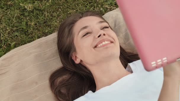 Top view medium close-up of happy beautiful Caucasian woman lying down on back on blanket over green lawn, using tablet computer in pink case, smiling - Footage, Video