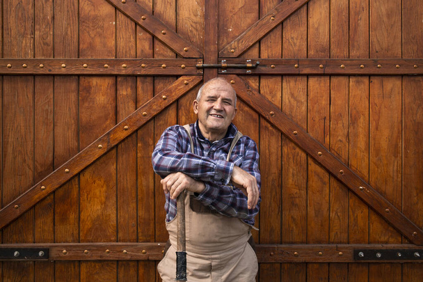 Portrét usměvavého farmáře stojícího u dveří dřevěné stodoly nebo potravinářské sýpky na farmě domácích zvířat. - Fotografie, Obrázek