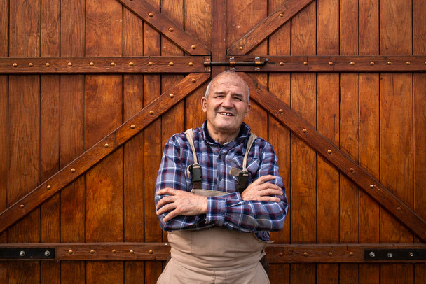 Portret van een lachende boer met gekruiste armen bij houten schuur of voedselgraandeuren op een boerderij voor huisdieren. - Foto, afbeelding