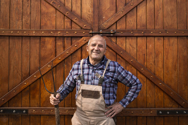 Ritratto di contadino laborioso e sorridente che tiene in mano un attrezzo da forcone e sta accanto alla stalla di legno o alle porte del granaio alimentare nell'allevamento di animali domestici. - Foto, immagini