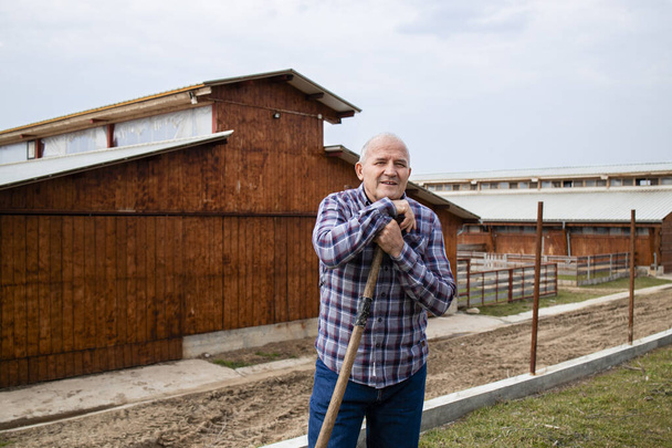 Portrait of successful farmer with pitchfork tool. In background wooden farmhouse. - Photo, Image