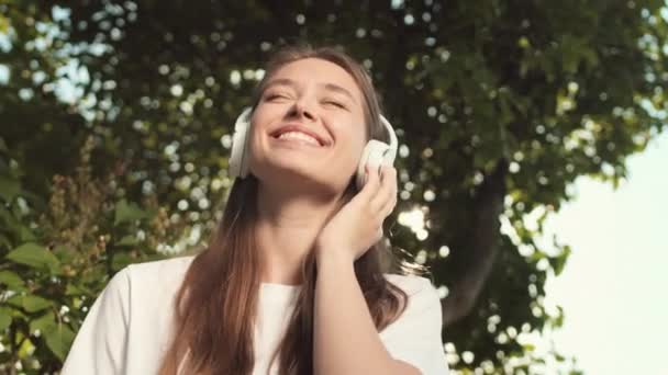 Nízký úhel hrudníku radostné mladé bělošky v ušních sluchátkách, poslouchající hudbu, pohybující se, stojící ve slunečném letním parku, usmívající se se zavřenýma očima - Záběry, video