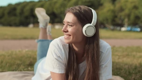 Track-in POV ευτυχισμένη όμορφη Καυκάσια γυναίκα φορώντας ακουστικά πάνω από το αυτί, κινούμενα πόδια, ξαπλωμένη στο στομάχι σε κουβέρτα πάνω από το πράσινο γκαζόν, χαμογελώντας και κοιτάζοντας την κάμερα - Πλάνα, βίντεο