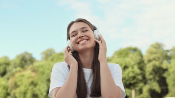 Borst-up POV van vrolijke jonge blanke vrouw dragen over-ear hoofdtelefoon, luisteren naar muziek, bewegen, staan in zonnige zomer park, glimlachen, dan zwaaien hand en kijken op camera - Video