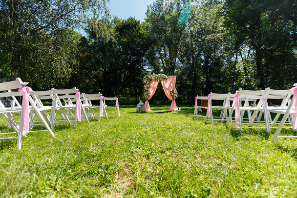 Área de la ceremonia de boda en el parque. Arco rectangular de madera, sillas blancas decoradas con flores, vegetación, pétalos, hojas y tul en color melocotón. Lindo, decoración rústica de moda - Foto, imagen