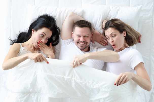 Man lying with two lovers in bed. Women looking under cover and wondering - Photo, Image