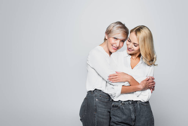 happy mother and daughter in jeans and white blouses embracing isolated on grey - Fotoğraf, Görsel