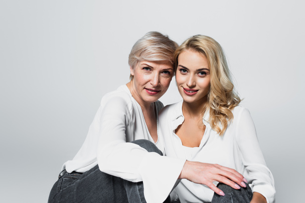 stylish mother and daughter smiling at camera while sitting isolated on grey - Foto, afbeelding