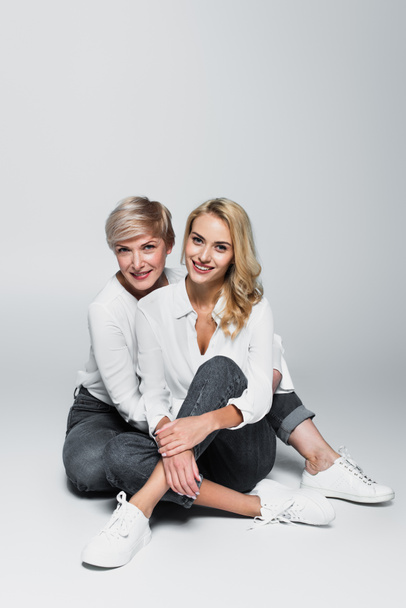 full length view of trendy mother and daughter smiling at camera while sitting on grey - Zdjęcie, obraz