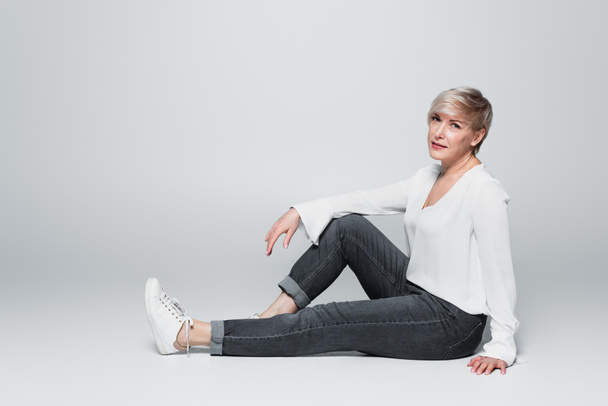 full length view of stylish, mature woman looking at camera while sitting on grey - Φωτογραφία, εικόνα