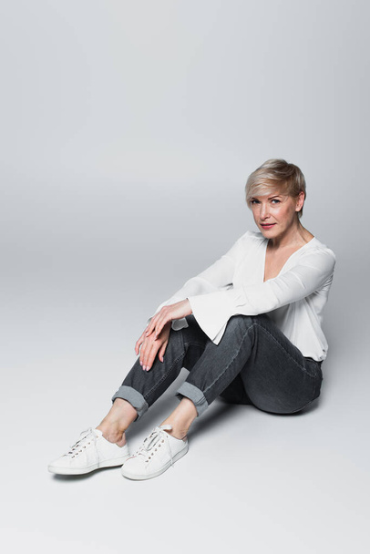 ash-blonde woman in white blouse and jeans looking at camera while sitting on grey - Foto, immagini