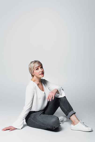 stylish, middle aged woman in white blouse and jeans sitting on grey - Fotoğraf, Görsel