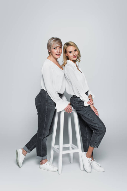 Вид в полный рост матери и дочери в джинсах и белых блузках, позирующих возле стульчика на сером фоне - Фото, изображение