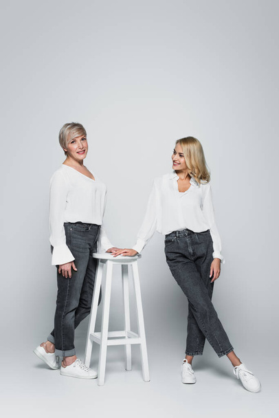 full length view of smiling middle aged woman posing near high chair and young daughter on grey - Foto, imagen