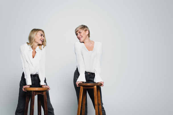 joyful mother and daughter smiling at each other while posing near high chairs on grey - Foto, imagen